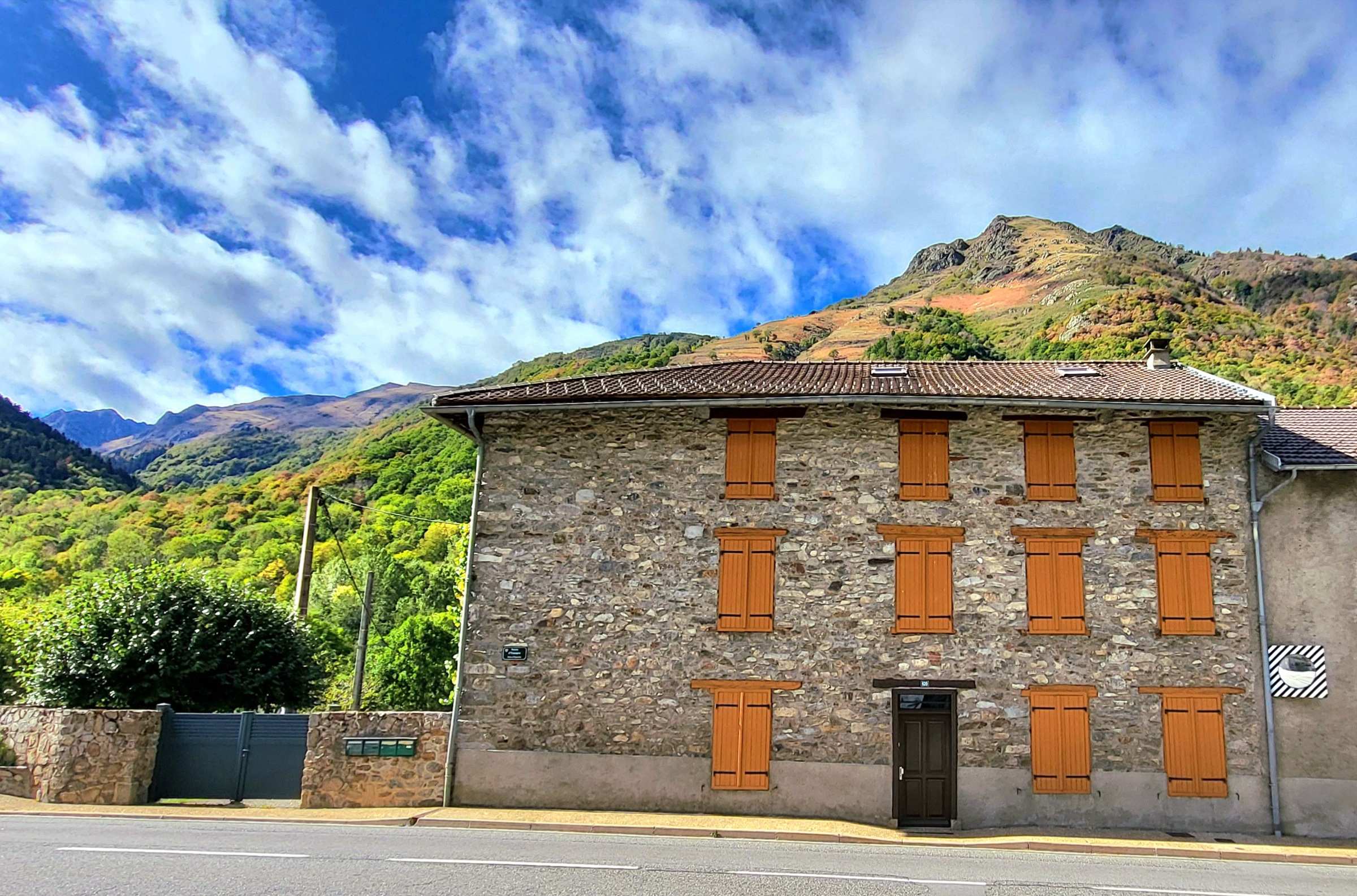La façade côté rue : pierre, volets orange et montagne en arrière-plan